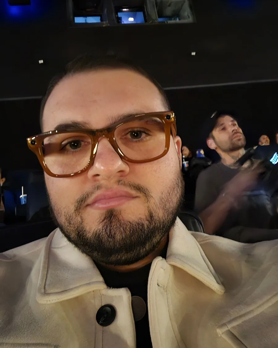 Selfie do Marco na poltrona da sala de cinema, de óculos e jaqueta clara, antes da sessão.