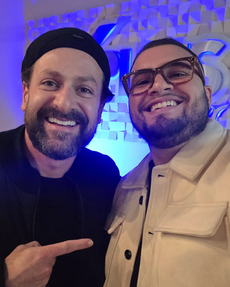 Selfie do Marco com um convidado no saguão do festival, os dois sorrindo diante de um painel iluminado de azul.