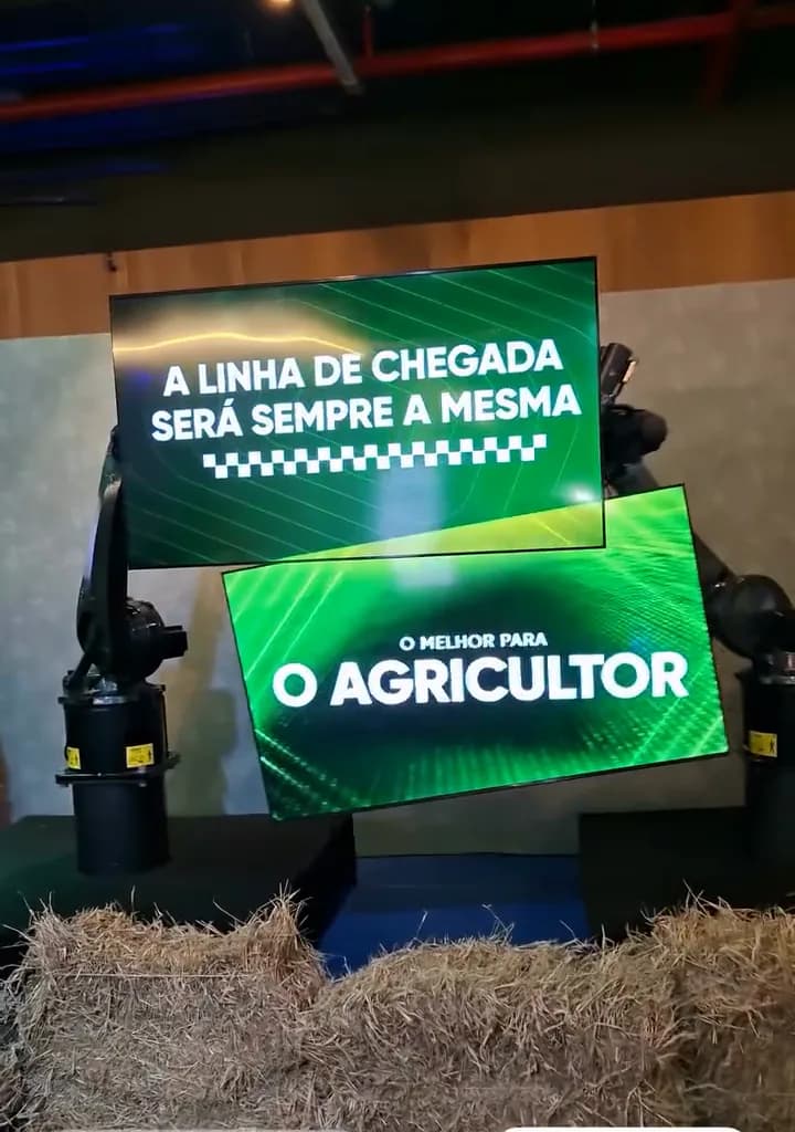 Registro do evento: dois monitores verdes presos a braços robóticos, com frases sobre o agricultor, sobre fardos de feno.