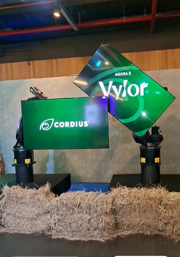 Registro do evento: os braços robóticos inclinam um monitor com o logo da Vylor ao lado de outro com o logo da Cordius.