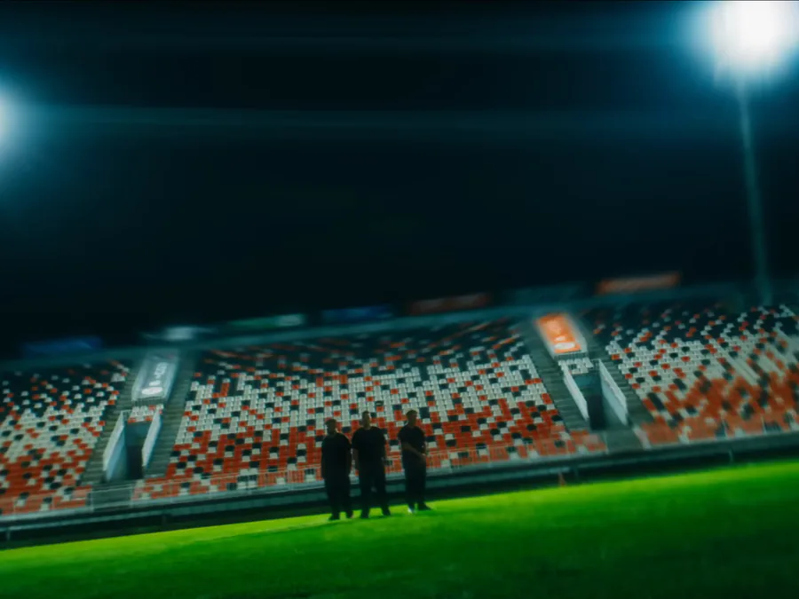 Quadro do anúncio de Rocket League: um estádio iluminado à noite, com três pessoas no gramado.