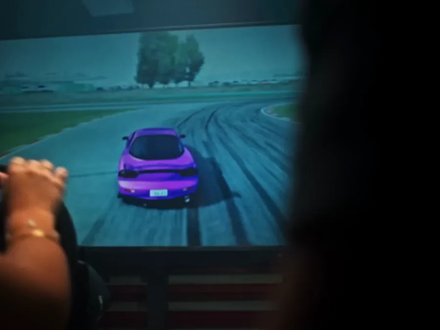 Quadro do filme: mãos no volante de um simulador, com um carro de drift na tela.
