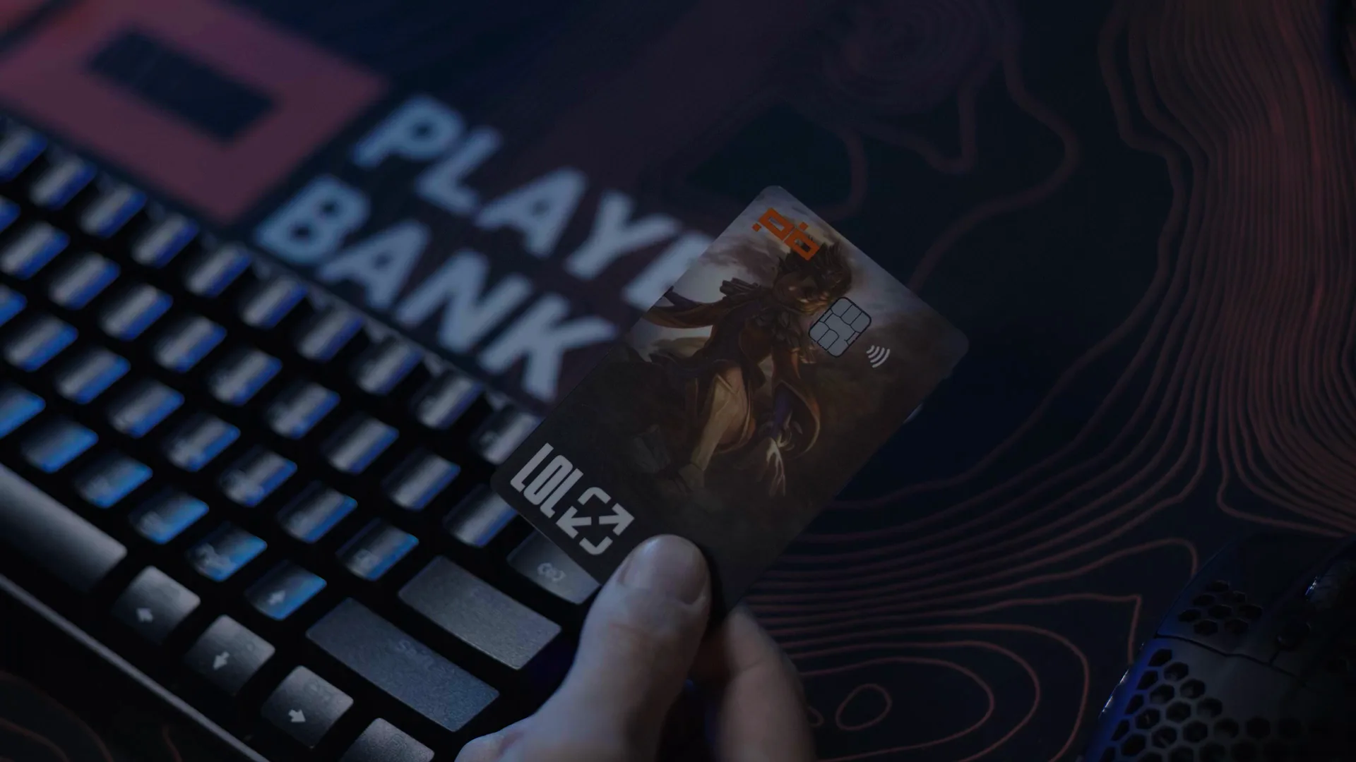 Uma mão segura o Cartão LoL sobre o teclado e o mousepad do Players Bank, quadro do filme.
