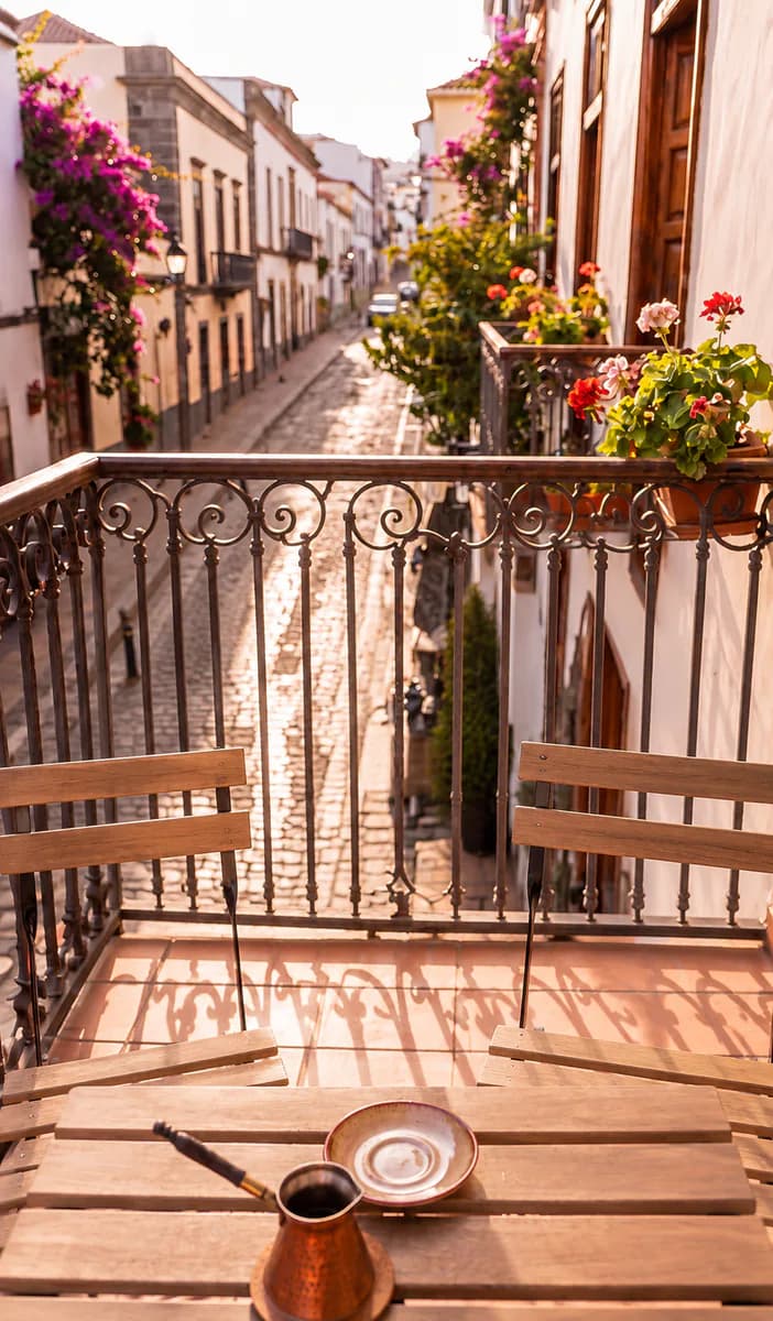 Varanda de ferro sobre uma rua de pedra com flores, com duas cadeiras e uma mesa.