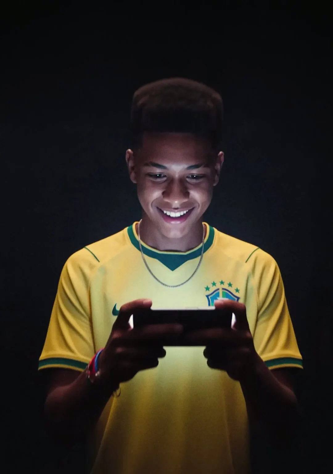 Jovem com a camisa da Seleção sorri olhando o celular no escuro.
