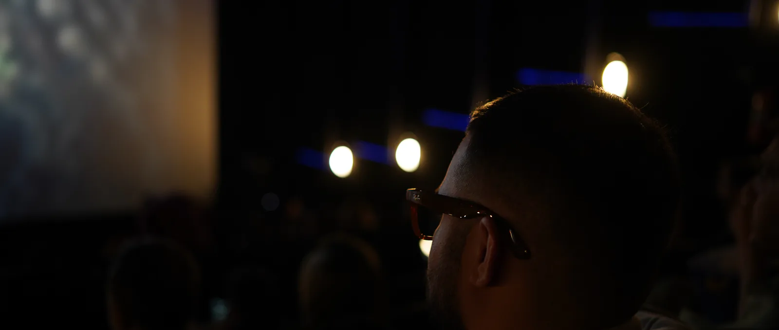 O Marco de perfil na plateia escura do cinema, de frente para a tela, na abertura da noite.